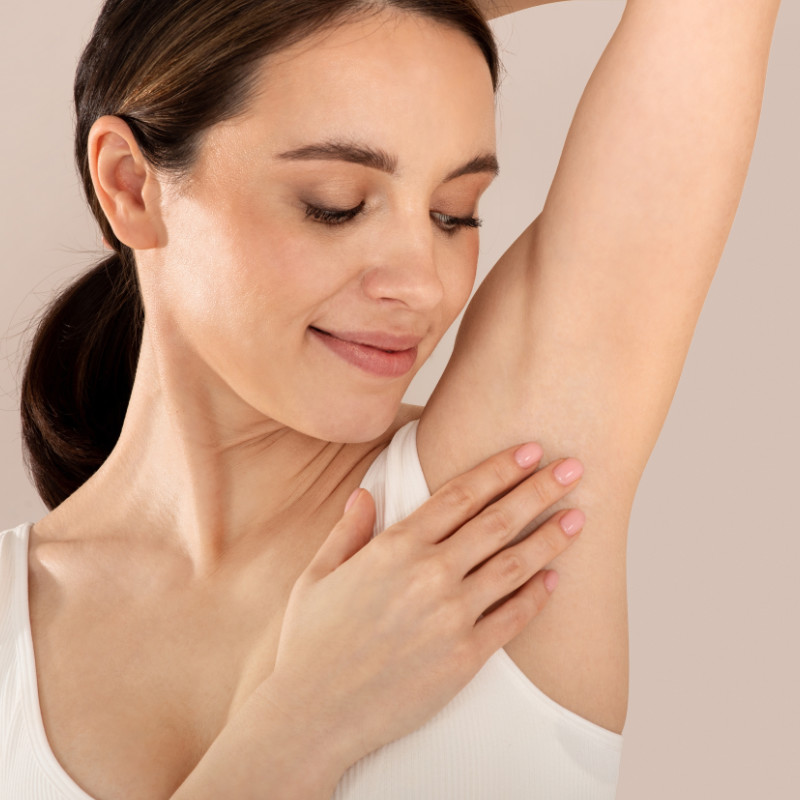 Woman in a white tank top smiling and touching the skin of her underarm while looking at it.