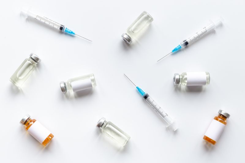 Vials filled with clear and amber liquids and three syringes with needles are arranged on a white background. The vials have blank white labels.